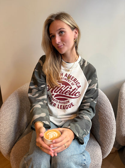 Dames sweater Athletic Camo met crème voorkant, bordeauxrode tekstprint en grijze camouflage mouwen, gecombineerd met een lichte jeans.