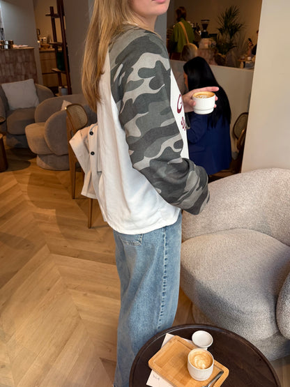 Dames sweater Athletic Camo met crème voorkant, bordeauxrode tekstprint en grijze camouflage mouwen, gecombineerd met een lichte jeans.
